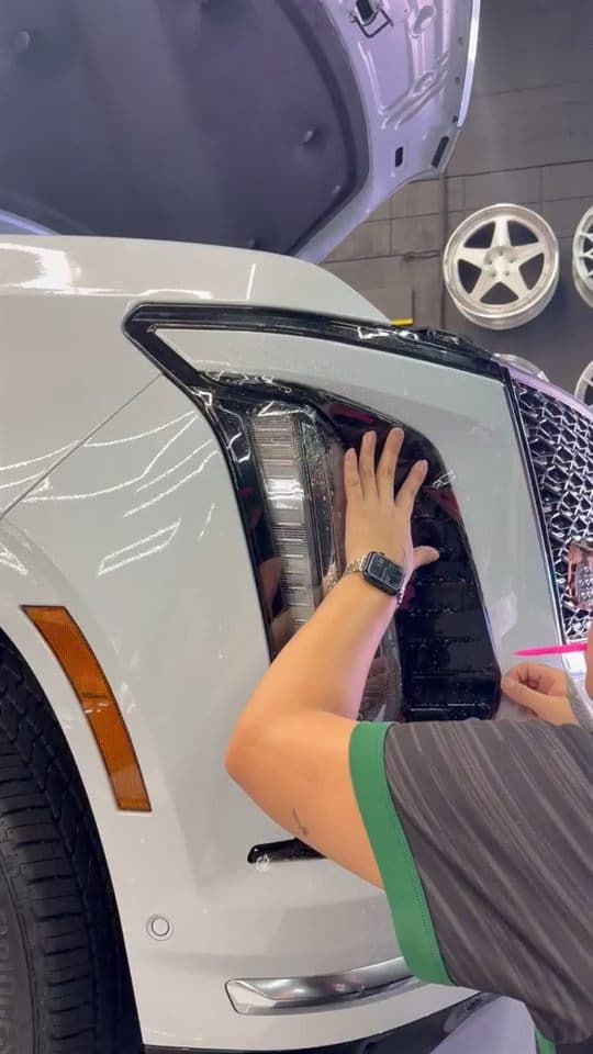 Hands squeegeeing paint protection film around the headlight of a white Cadillac Escalade, wheels on display behind